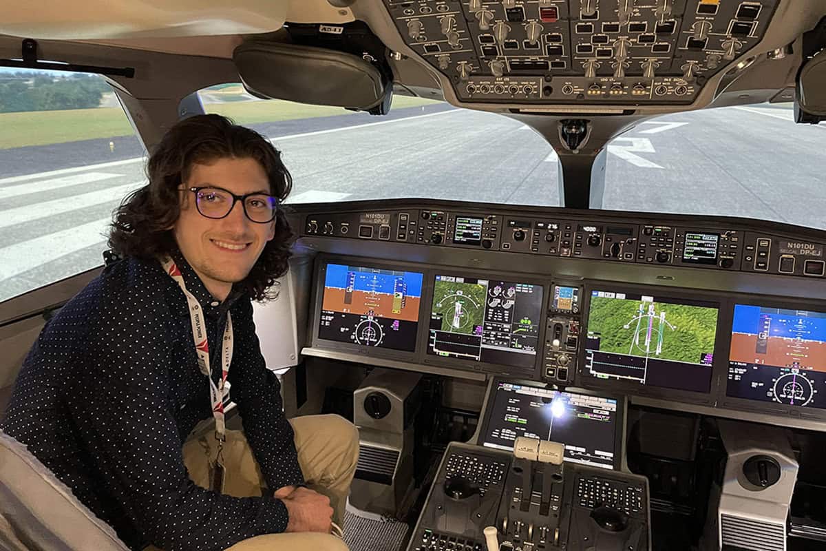 Sinan sits in an airplane engine room smiling at the camera with a visitor lanyard around his neck
